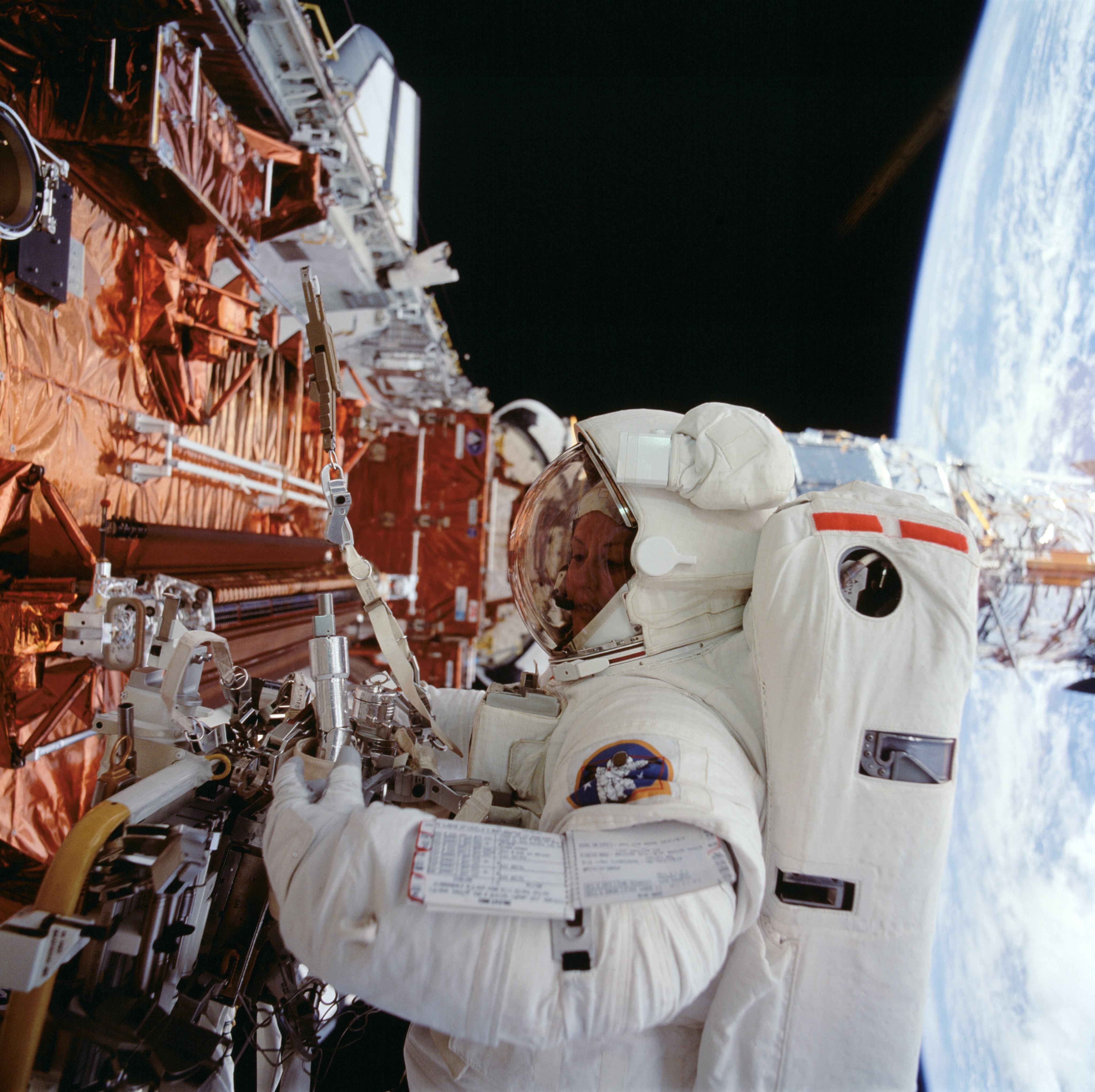Astronaut Kathryn Thornton works with instruments while on a spacewalk. A small part of Earth is visible behind her on the right.)