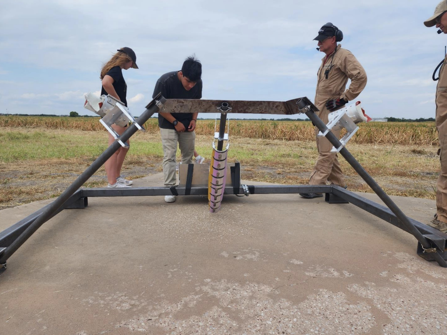 Three people stand at a rig with three angled beams. Left and right beams have equipment attached. Middle vertical beam has a pink/yellow attachment. 