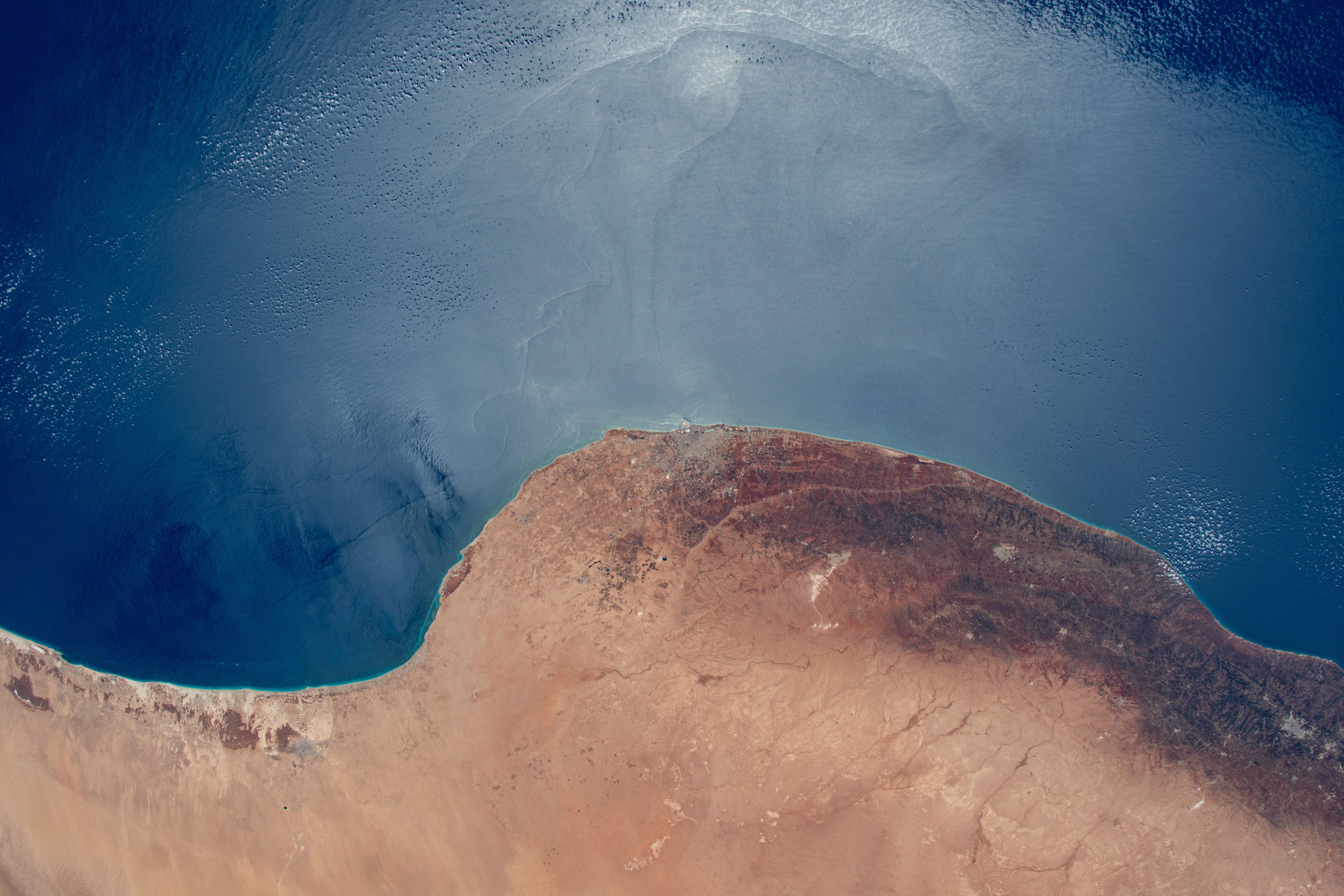 The sun's glint and the deep blue waters of the Mediterranean Sea stand in stark contrast to the desert coast of Libya in this photograph from the International Space Station as it orbited 260 miles above. Benghazi, a city of 1.2 million people, is at center in the African nation's arid region in between its desert and temperate climates.