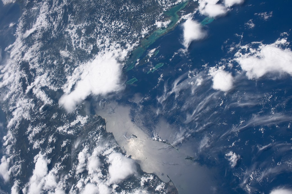 The Sun casts a glass-like reflection on the Atlantic Ocean off the coast of Honduras. Near the top of the image, groups of teal waters off the coast of Belize peek through the clouds as the International Space Station soared nearly 260 miles above.