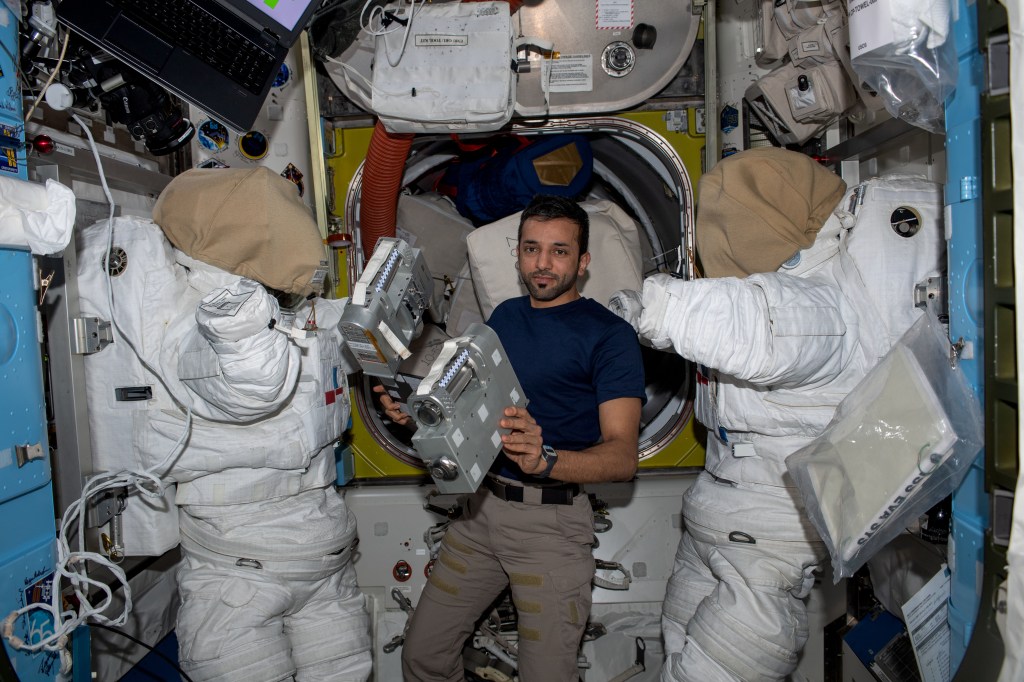 UAE (United Arab Emirates) astronaut and Expedition 69 Flight Engineer Sultan Alneyadi is pictured in between a pair of spacesuits while working on hardware maintenance inside the International Space Station's Quest airlock.