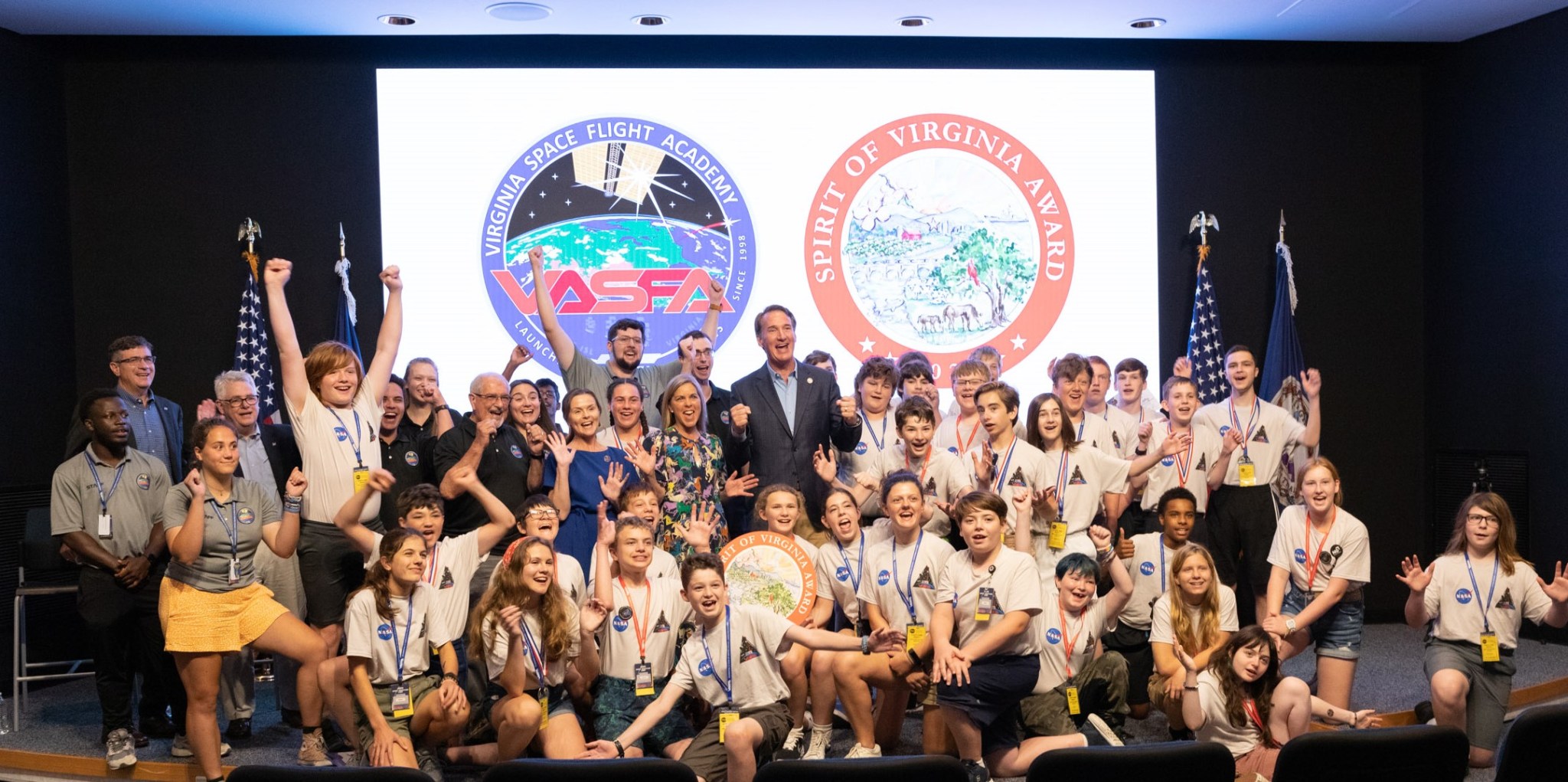 a group of 40 people, mostly youth celebrate with raised arms and large smiles on a stage in front of a screen showing a Virginia Space Flight Academy logo and a Spirit of Virginia logo.