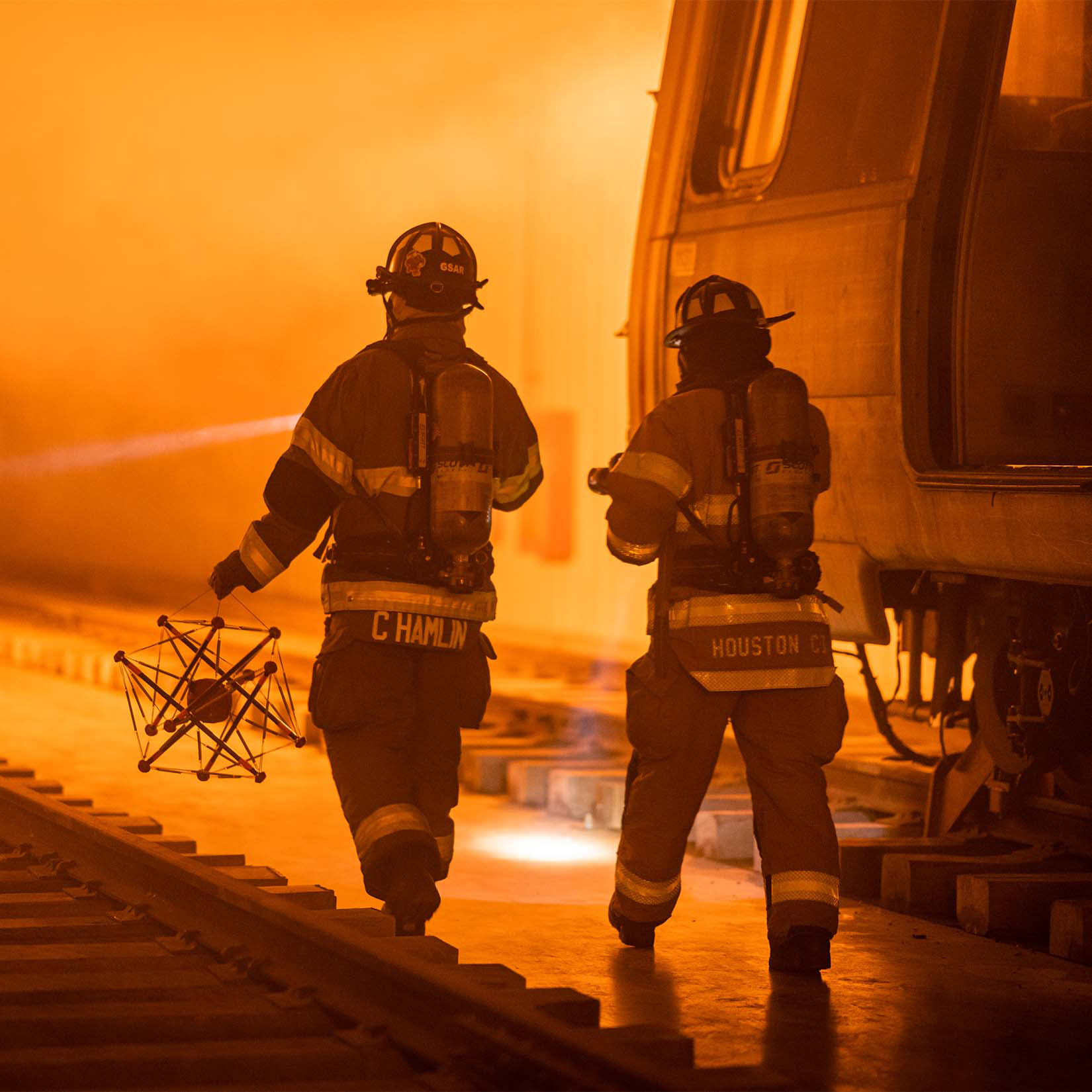 Inside of an underground subway station, two firefighters carry a robot mounted inside of a trapezoid-shaped wireframe toward a blazing fire. There is a subway car to the firefighters' right, highlighted by the red, yellow, and orange hue of a fire in the background. The robot helps firefighters and other first responders protect their lives and the lives of others by investigating hazardous situations.