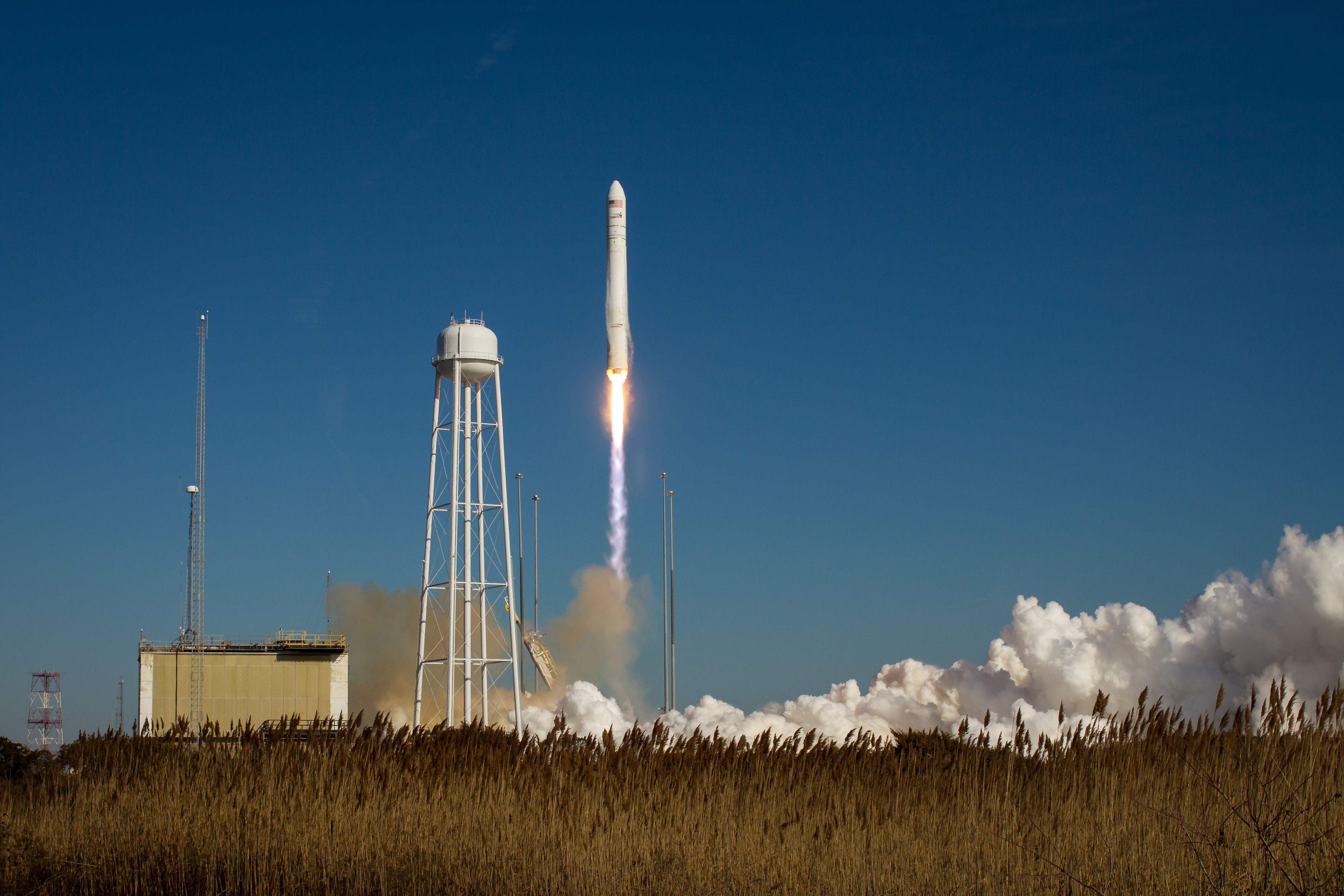 A white rocket launches upward, leaving a short trail of vapor behind it. The flame at the bottom of the rocket is a bright spot near the center of the photo. To the left of the rocket is a white tower and other structures. Thick plumes of white smoke frame the field in the foreground.