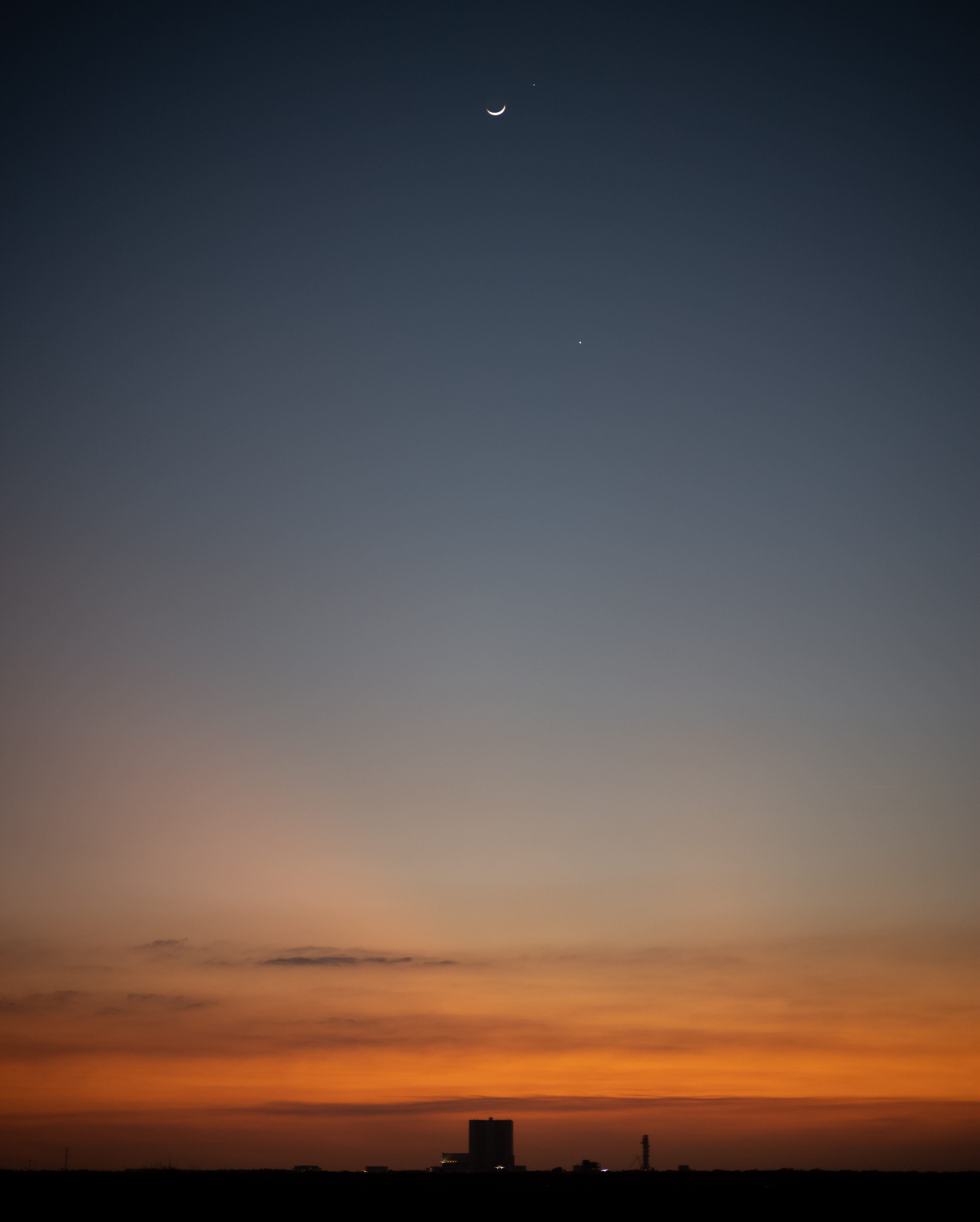 The sky shifts from a deep blue to burnt orange (top to bottom). The crescent moon is at the top of the image, with Jupiter to its right and Venus below it. The silhouette of the Vehicle Assembly Building at NASA's Kennedy Space Center stands out against the orange sky.