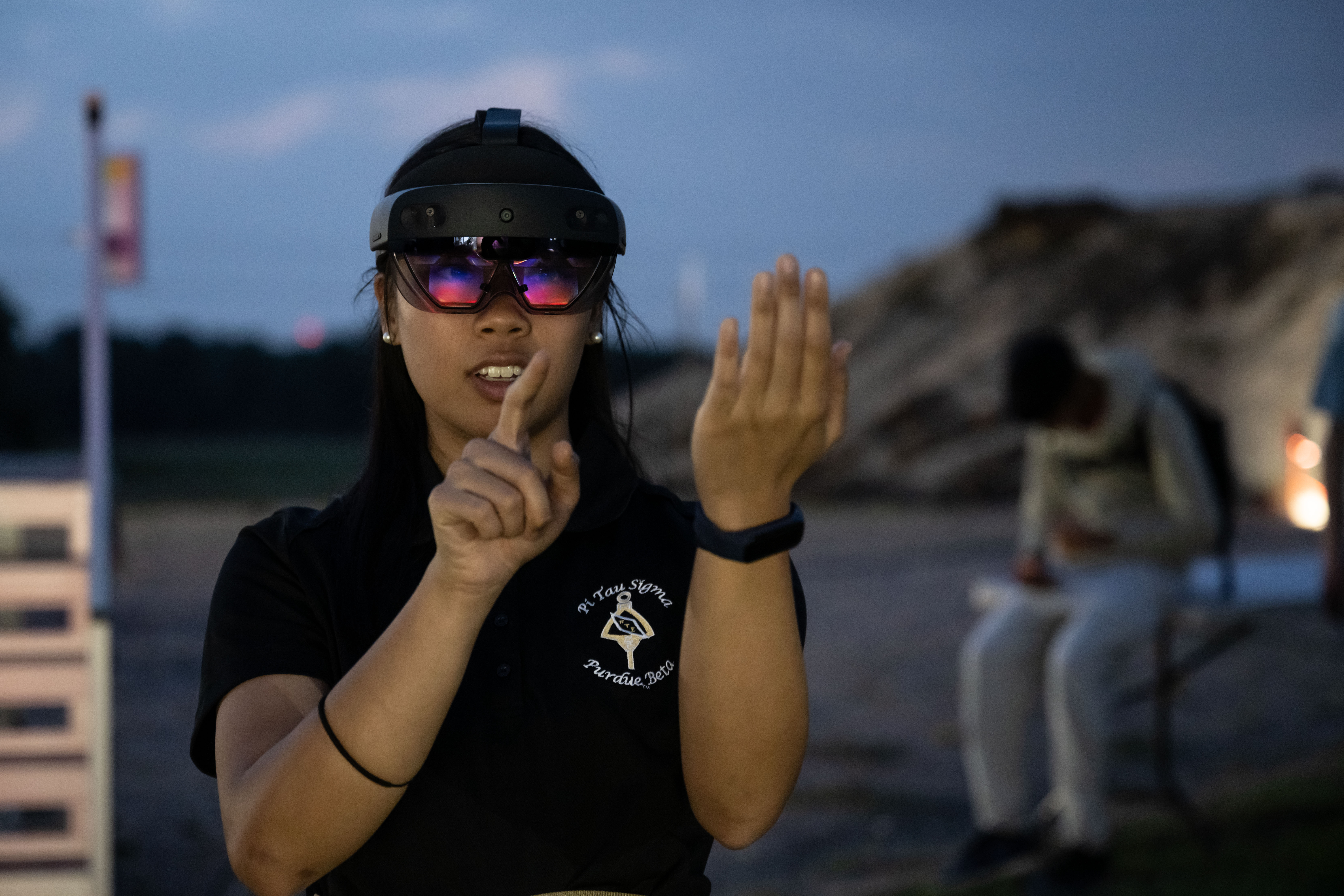 A NASA intern wearing a dark-colored polo shirt stands in a rocky outcrop at the aptly named “Rockyard” at NASA’s Johnson Space Center in Texas as the sun sets. In the background, another intern sits on a camping chair and the sky is light greyish blue of early dusk.