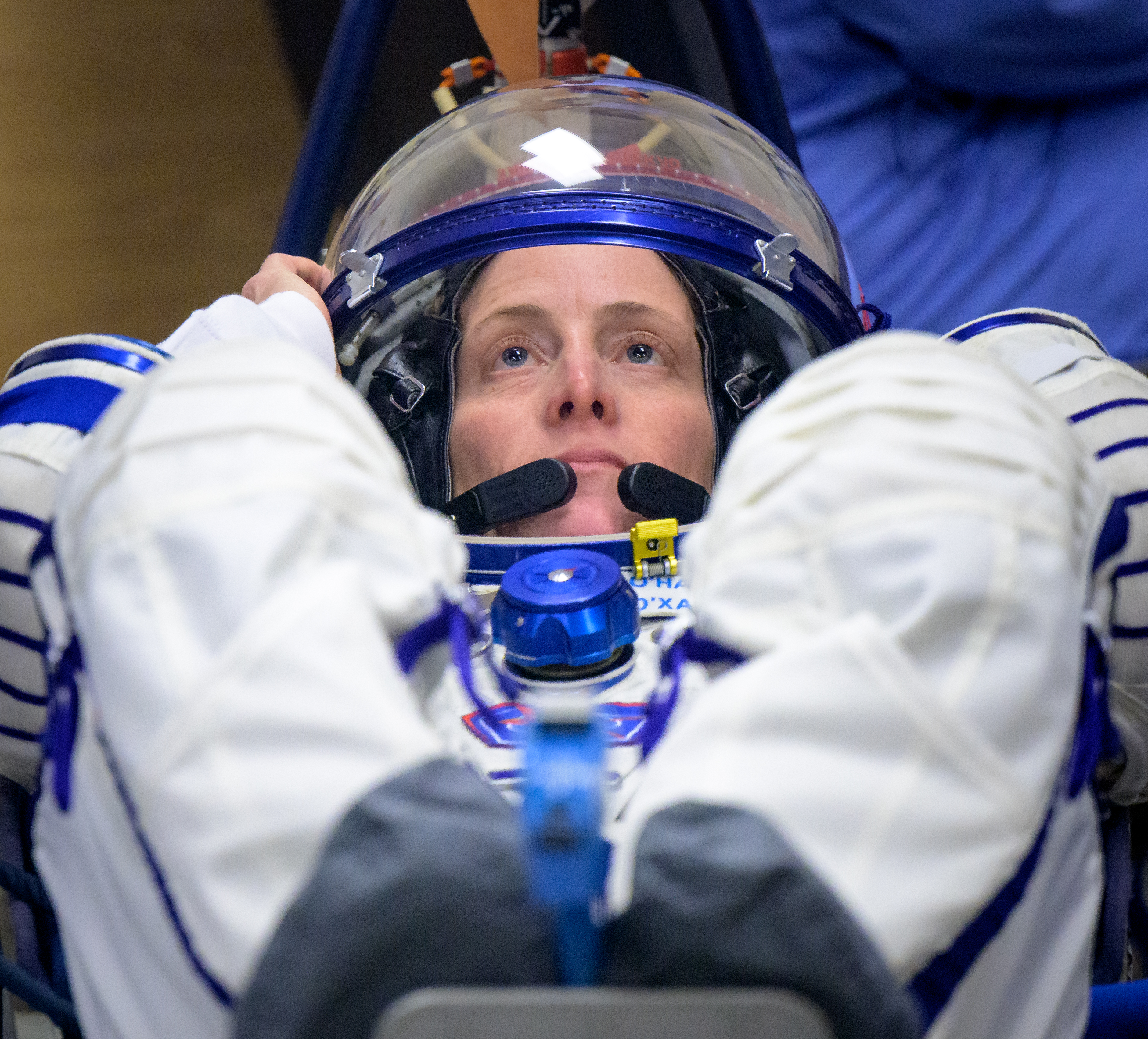Astronaut Loral O'Hara looks upward while in a reclined position. Her feet are out of focus in the foreground. Her hand rests on the blue rim of her helmet. She wears a white and blue spacesuit.
