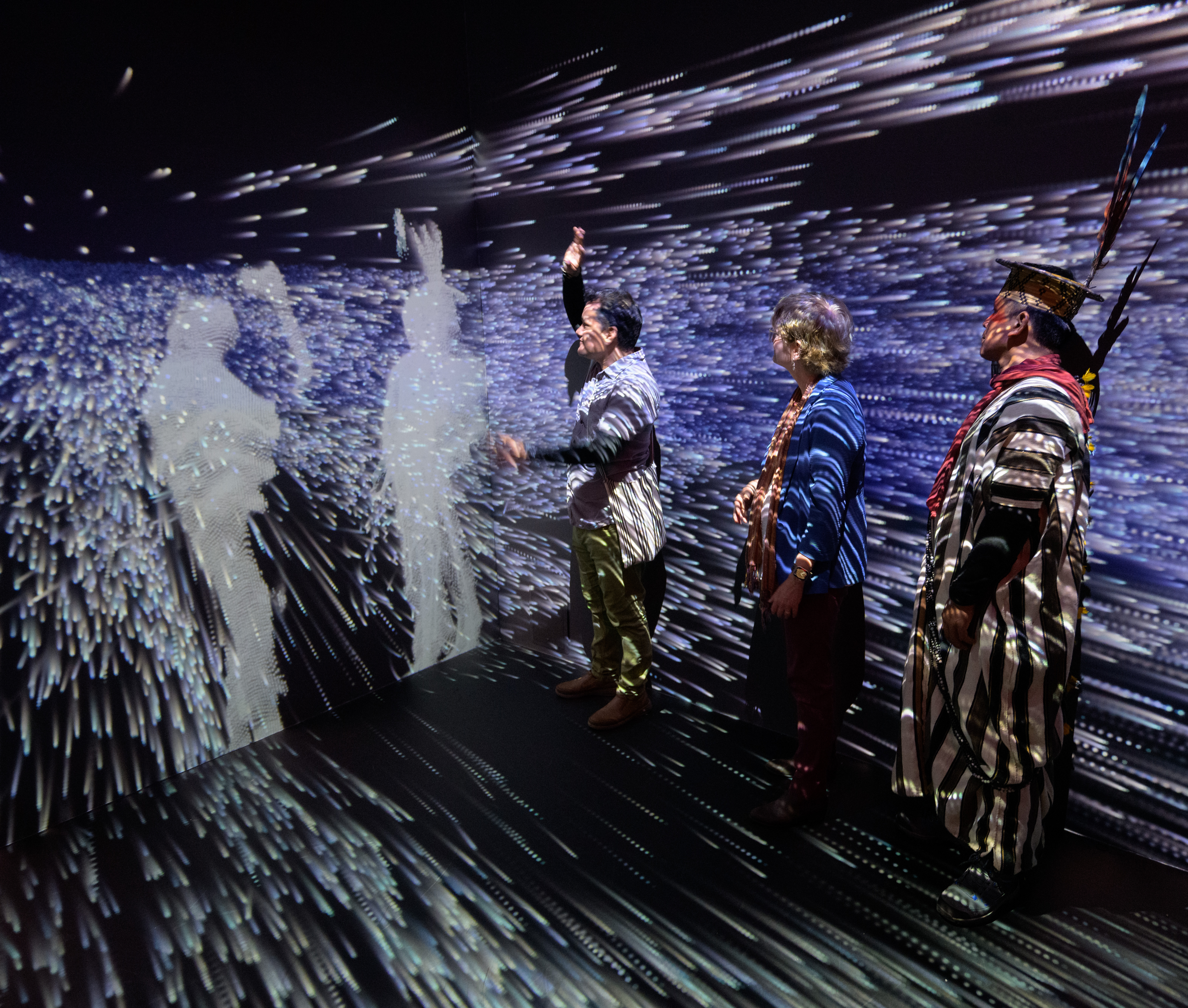 Amazonian leaders stand in the small "Space for Earth" immersive installation room, with tiny white dots of light representing atmospheric rivers flowing all around them on the walls and floor.