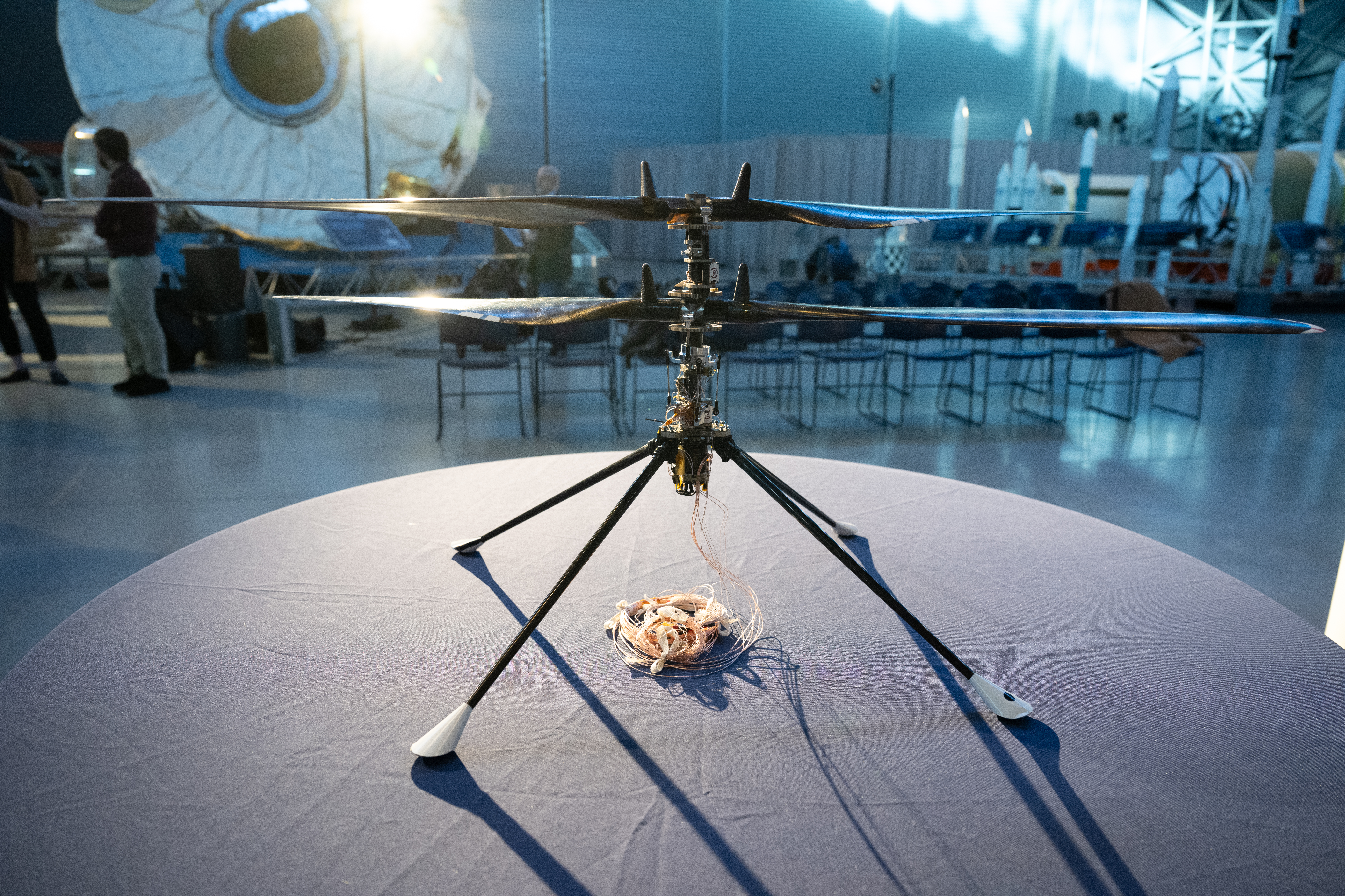 The Ingenuity Mars Helicopter prototype sits on a round, gray surface. Sunlight glints off of one of its blades. The blades are stacked vertically on a thin column. Four thin legs with white tips are attached to the column. A bundle of wires protrudes from the helicopter and rests on the table.