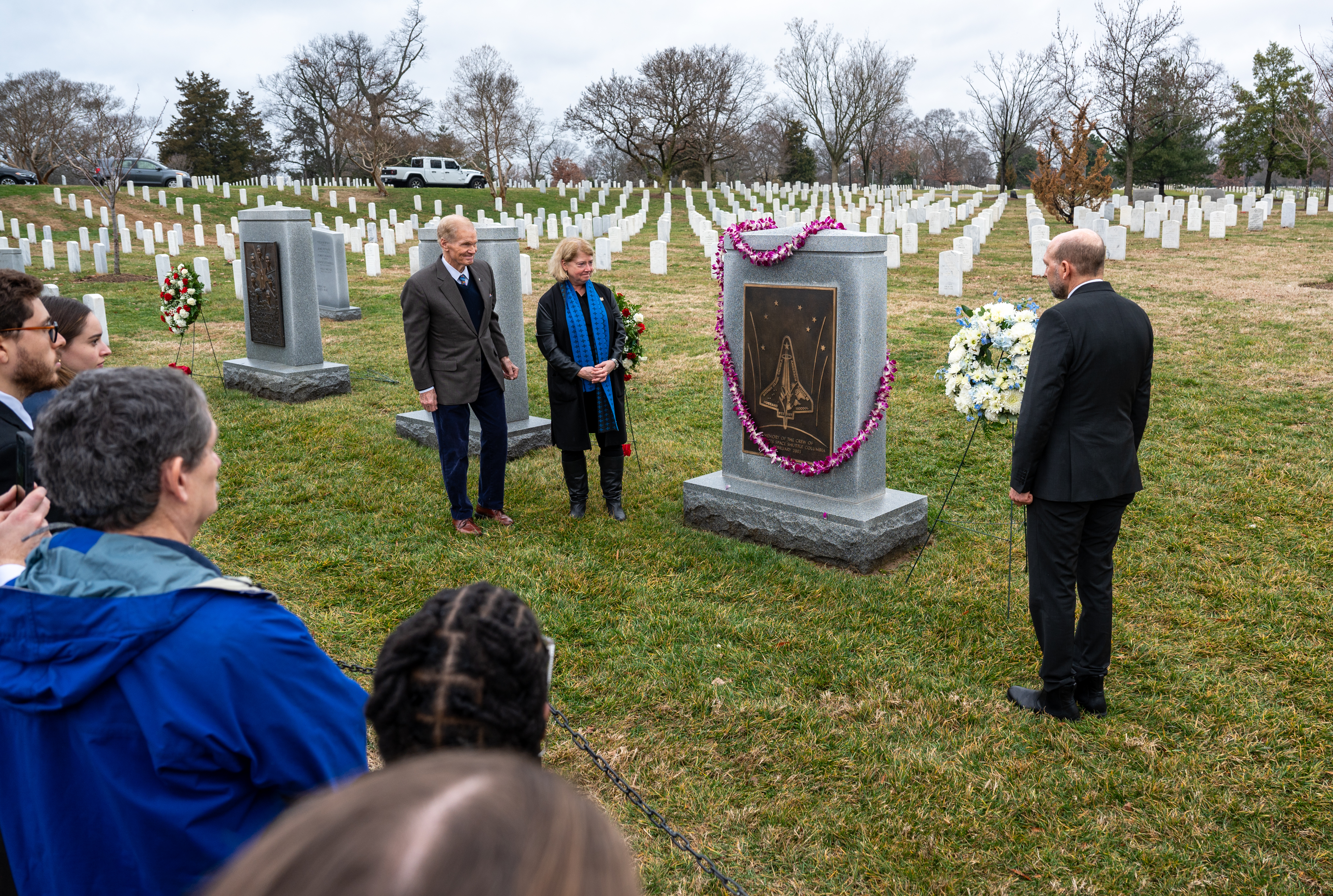 NASA Administrator Bill Nelson (far left), NASA Deputy Administrator Pam Melroy (left), and Deputy Chief of Mission for the Embassy of Israel Eliav Benjamin (right) stand on opposite sides of the Space Shuttle Columbia Memorial at Arlington National Cemetary. They are all dressed in dark clothing and look at the memorial. The memorial is a rectangular dark gray stone with a large bronze plaque on it; it has a large pink flower wreath draped on it. White headstones line the background. In the left foreground, NASA employees look on.