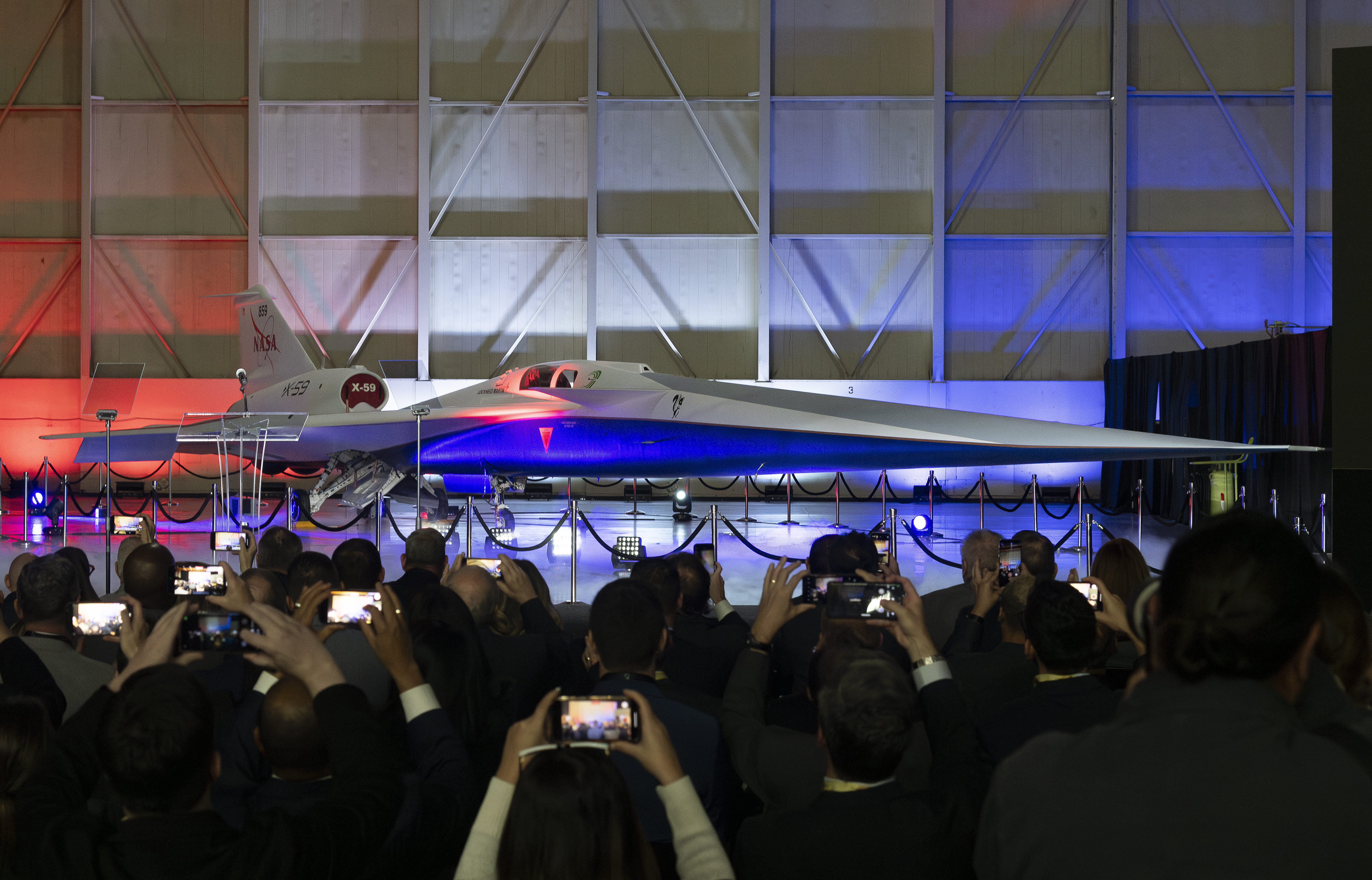 Members of an audience lift their phones to snap a photo of the recently revealed X-59 aircraft. The plane's long, sharp nose appears to jut out towards the audience.