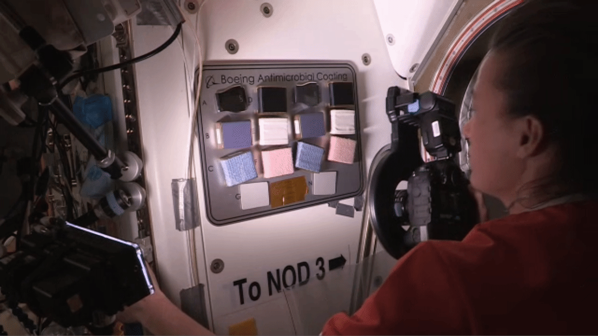 McArthur, wearing a red shirt, steadies herself with her left hand and aims a camera in her right hand at a rectangular panel on a wall of the space station labeled Boeing Antimicrobial Coating with 15 squares of different materials and colors.