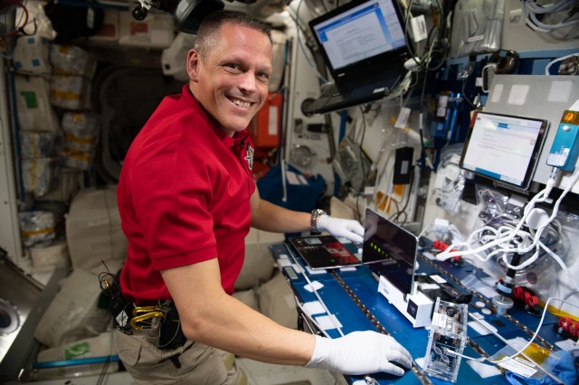 Bob Hines, wearing a red work polo and white gloves, smiles at the camera from his position at a workstation with many screens and wires aboard the International Space Station.