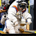An Axiom Space engineer kneels down to collect simulated lunar samples using a geology tool while wearing the AxEMU (Axiom Extravehicular Mobility Unit) spacesuit during testing at NASA’s Johnson Space Center.