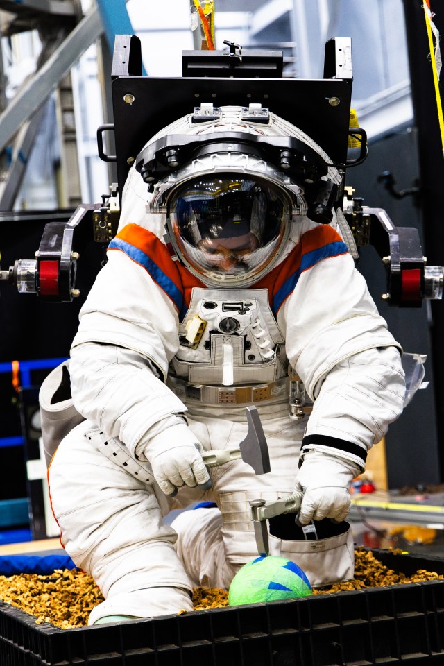 An Axiom Space engineer uses a hammer and chisel to chip off simulated lunar rocks while wearing the AxEMU (Axiom Extravehicular Mobility Unit) spacesuit during testing at NASA’s Johnson Space Center.