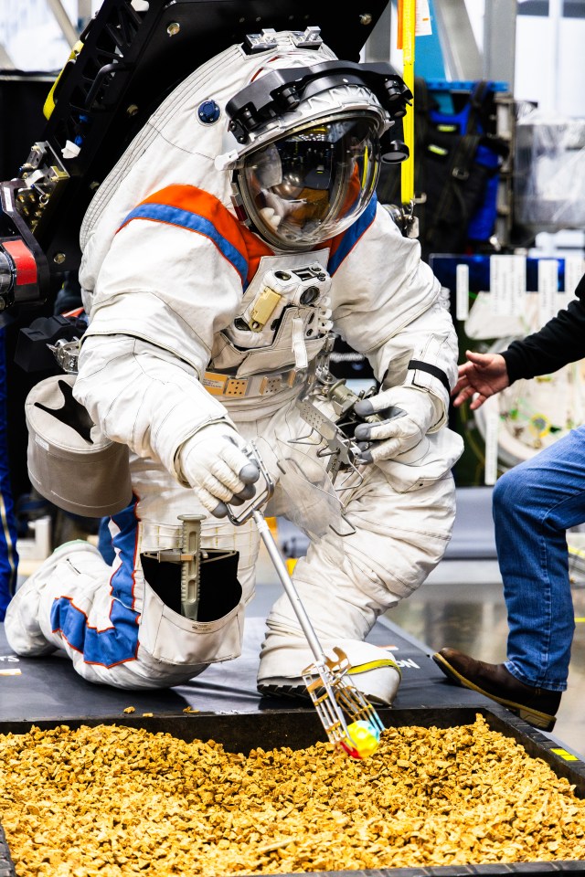 An Axiom Space engineer uses tongs to pick up a simulated lunar rock while wearing the AxEMU (Axiom Extravehicular Mobility Unit) spacesuit during testing at NASA’s Johnson Space Center.