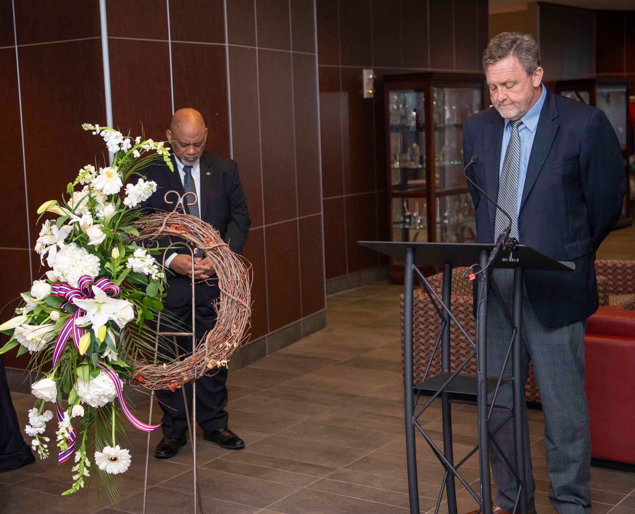 A man stands behind a lectern. A memorial wreath is displayed next to him.