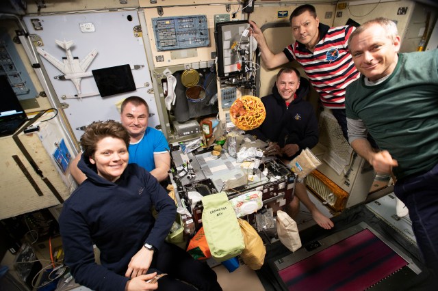 Five of six Expedition 59 crewmembers gather inside Russia's Zvezda service module for dinner as a pizza loaded with toppings floats above the galley. From left are Flight Engineers Anne McClain, Alexey Ovchinin and Nick Hague; Commander Oleg Kononenko and Flight Engineer David Saint-Jacques.
