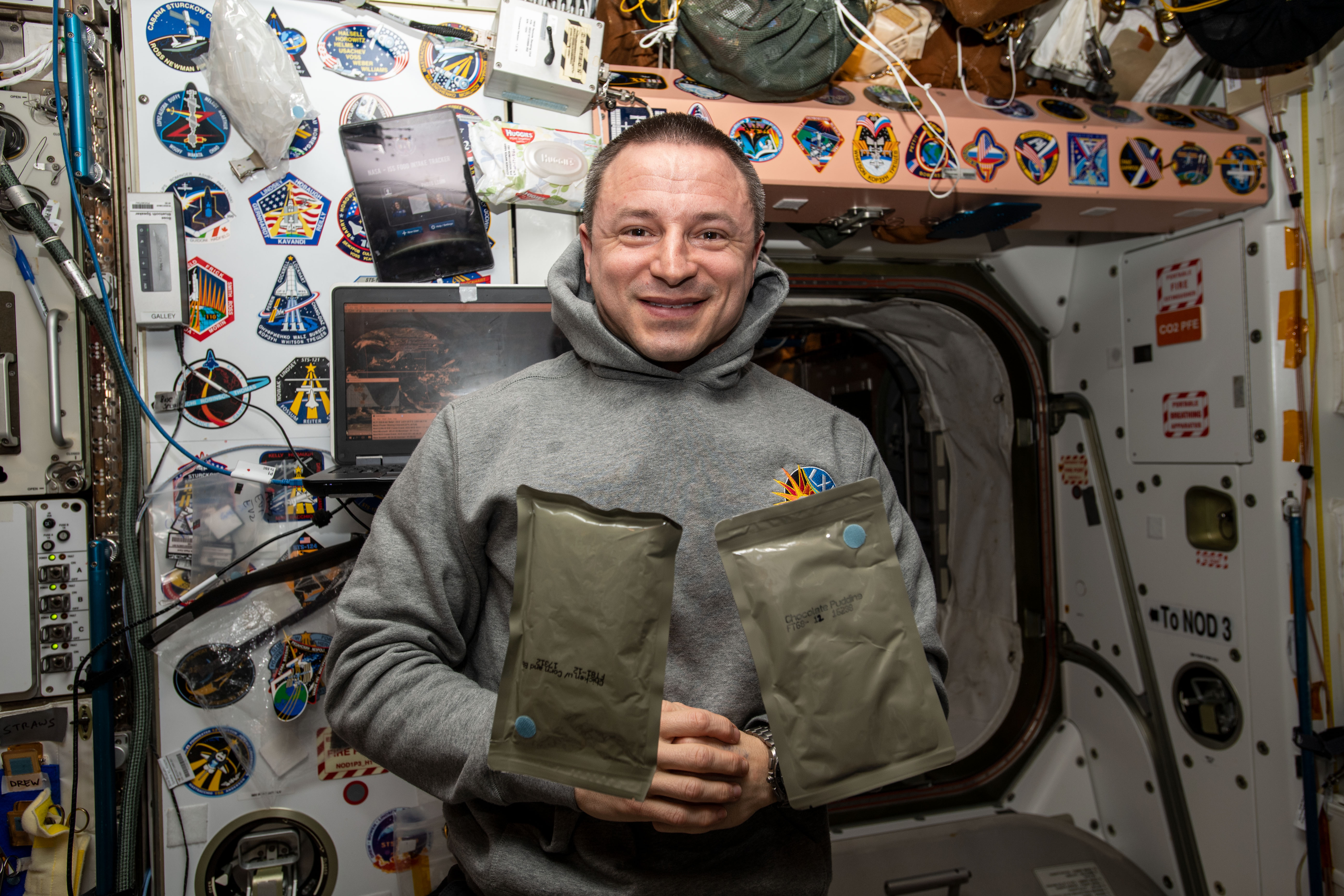 NASA astronaut and Expedition 61 Flight Engineer Andrew Morgan poses with two food packets, chicken with cornbread and chocolate pudding, during dinner time aboard the International Space Station's Unity module.