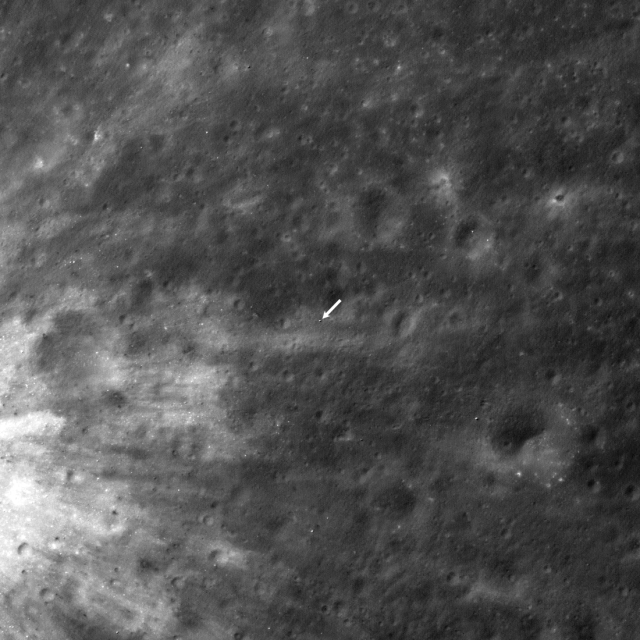 A black and white image of the surface of the moon taken from the Lunar Reconnaissance Orbiter. A white arrow in the center of the image points to the location of the SLIM lander.