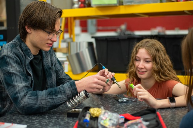 Sitting at a lab table, young man holds small component and young woman holds soldering iron.