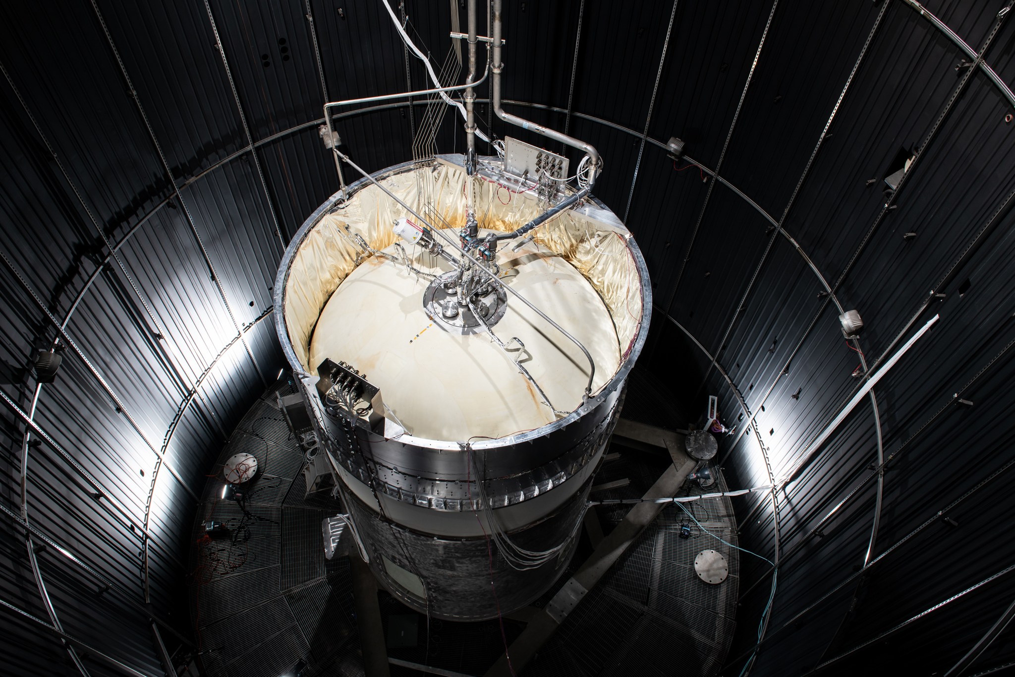 A view looking down on the test version of a cryogenic propellant tank, which stands vertically inside of NASA’s large vacuum chamber at the Neil Armstrong Test Facility in Sandusky, Ohio. The cream-colored top of the tank is illuminated by lights but the grey walls of the vacuum chamber that surround it are shadowed.