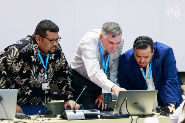 NASA Glenn Research Center’s Spectrum Manager Wayne White, center, stands and points at a computer screen at a long table with other computers and microphones. Two other delegates stand by him and view the screen.