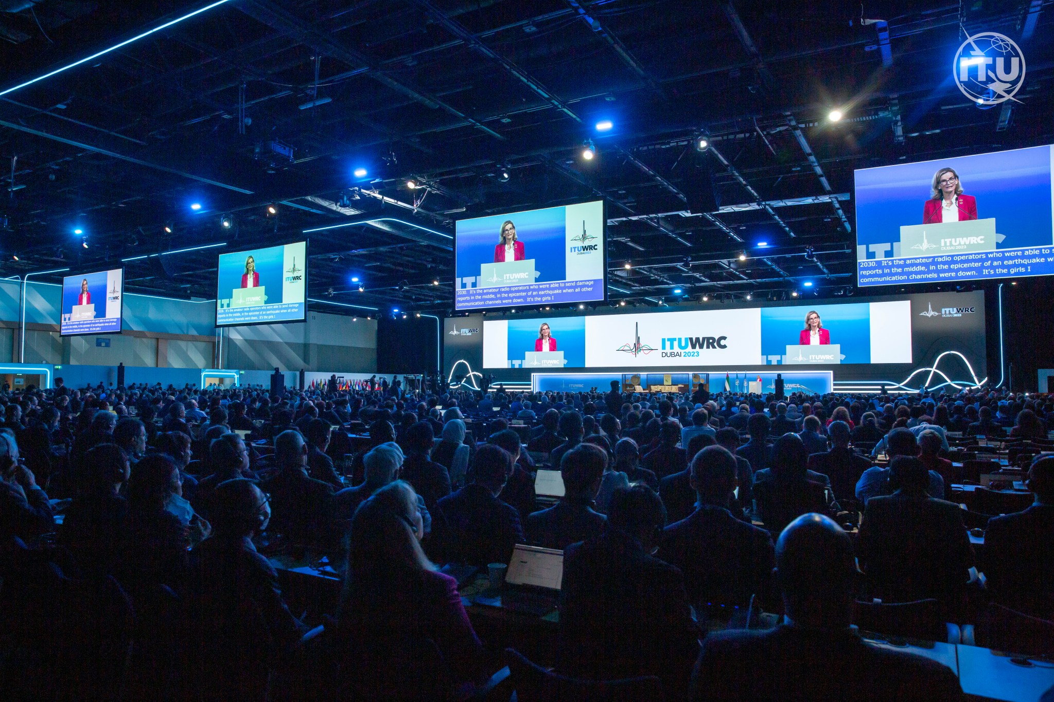 A woman speaks at a podium in front of a room of hundreds of delegates. Several large screens are placed around the stage.