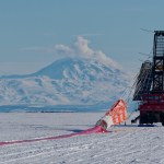 A large scientific observatory with reflective solar panels is suspended from a crane while connected to an orange parachute that is laid on the snow-covered ground. A mountain covered in snow is in the distance.