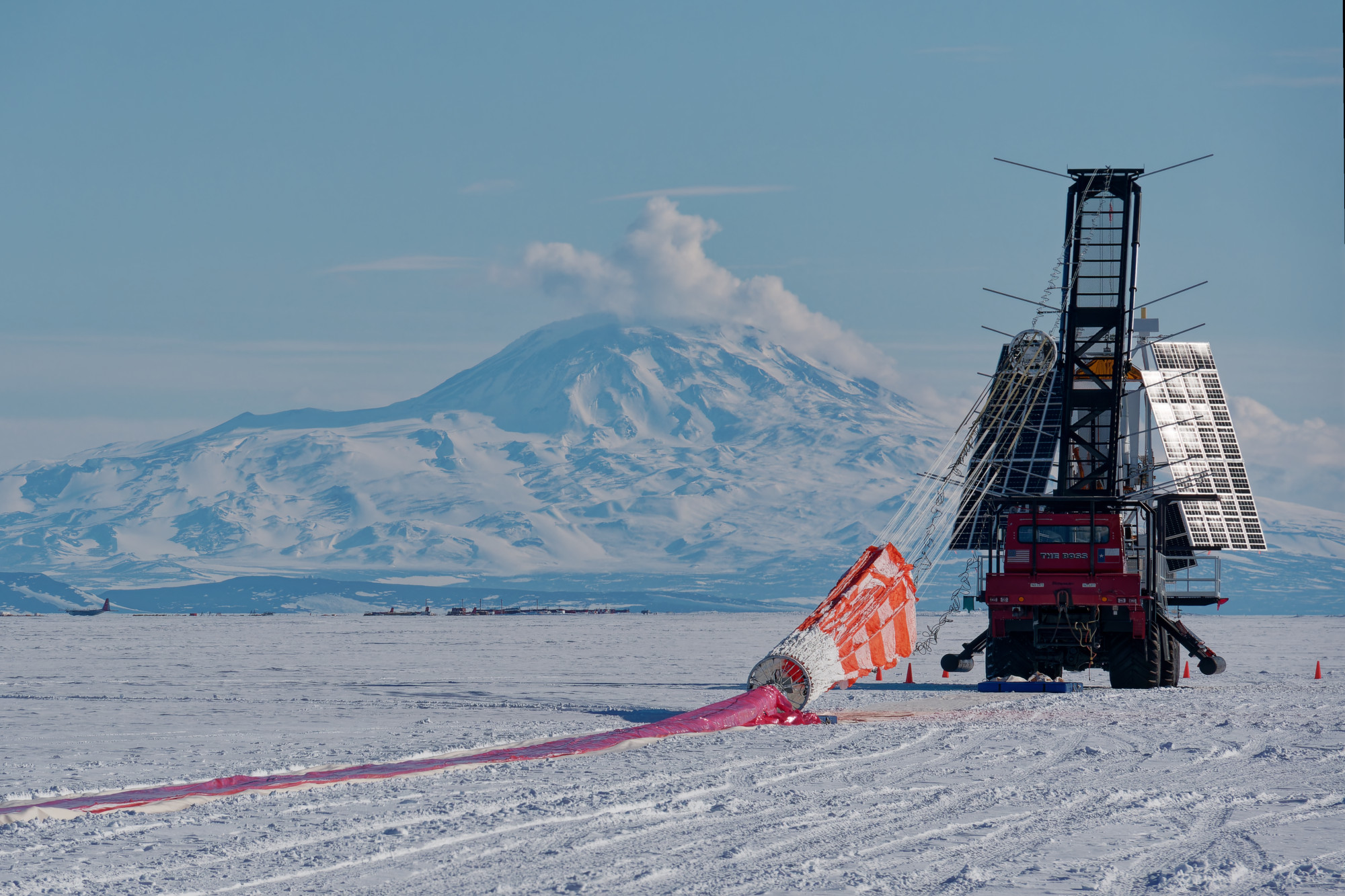 A large scientific observatory with reflective solar panels is suspended from a crane while connected to an orange parachute that is laid on the snow-covered ground. A mountain covered in snow is in the distance.