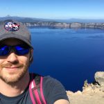 Joshua Schlieder, a man with a short brown beard, smiles and holds the camera up in a selfie outdoors near Crater Lake, Oregon. Joshua wears blue reflective sunglasses, a gray baseball cap with the NASA logo, and a navy T-shirt, and carries a backpack with pink straps. He takes up part of the left side of the image, with the still, deep blue lake visible in the background. Tan rocks and dirt are visible in the foreground, and the lake's rocky opposite shore is visible in the distance, with blue mountains on the horizon. It is a bright sunny day and the sky is cloudless and blue.