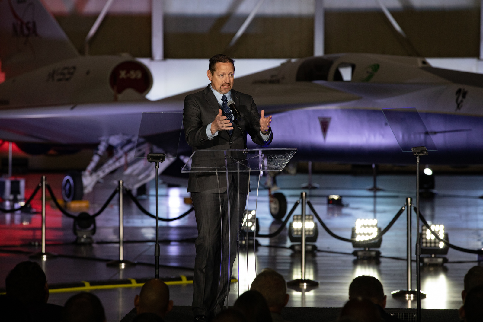 A man speaks at a podium. In the immediate background, the X-59 sits, newly painted in its red, white, blue and gold livery.