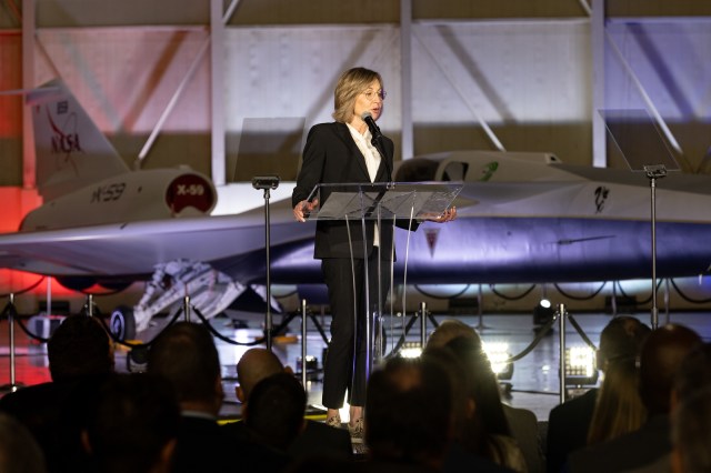 A woman speaks at a podium. In the immediate background, the X-59 sits, newly painted in its red, white, blue and gold livery.