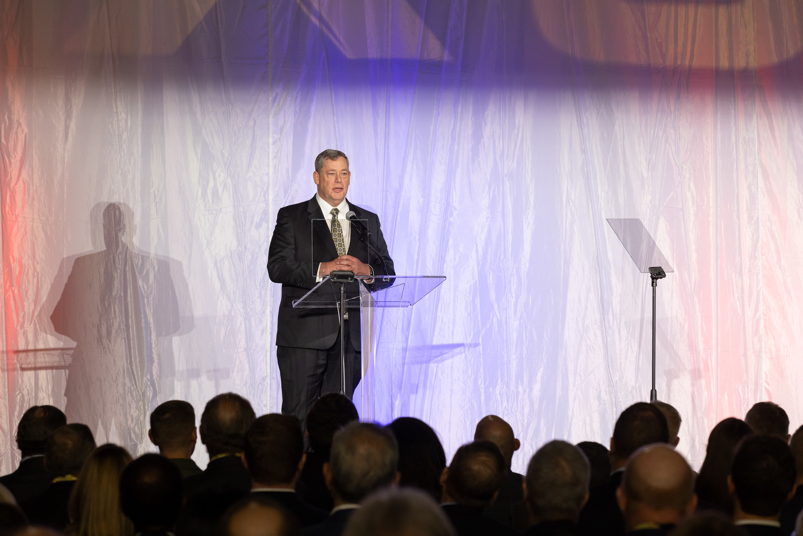 A man speaks at a podium. In the immediate background, the X-59 sits unseen behind a large curtain, moments away from its public unveiling.