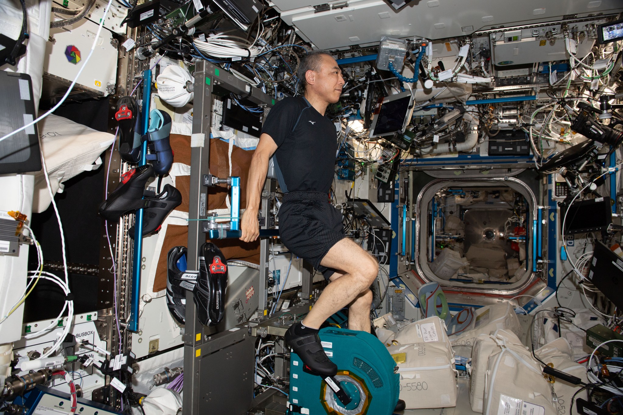 Furukawa wears a black shirt, shorts, and shoes. He holds on to a bar behind him and has his feet on the pedals on CEVIS, a teal boxy device about the size of a sofa cushion. Three pairs of shoes hang from bars to his right and the walls around him are covered in equipment, laptops, cords, hoses, and storage bags.