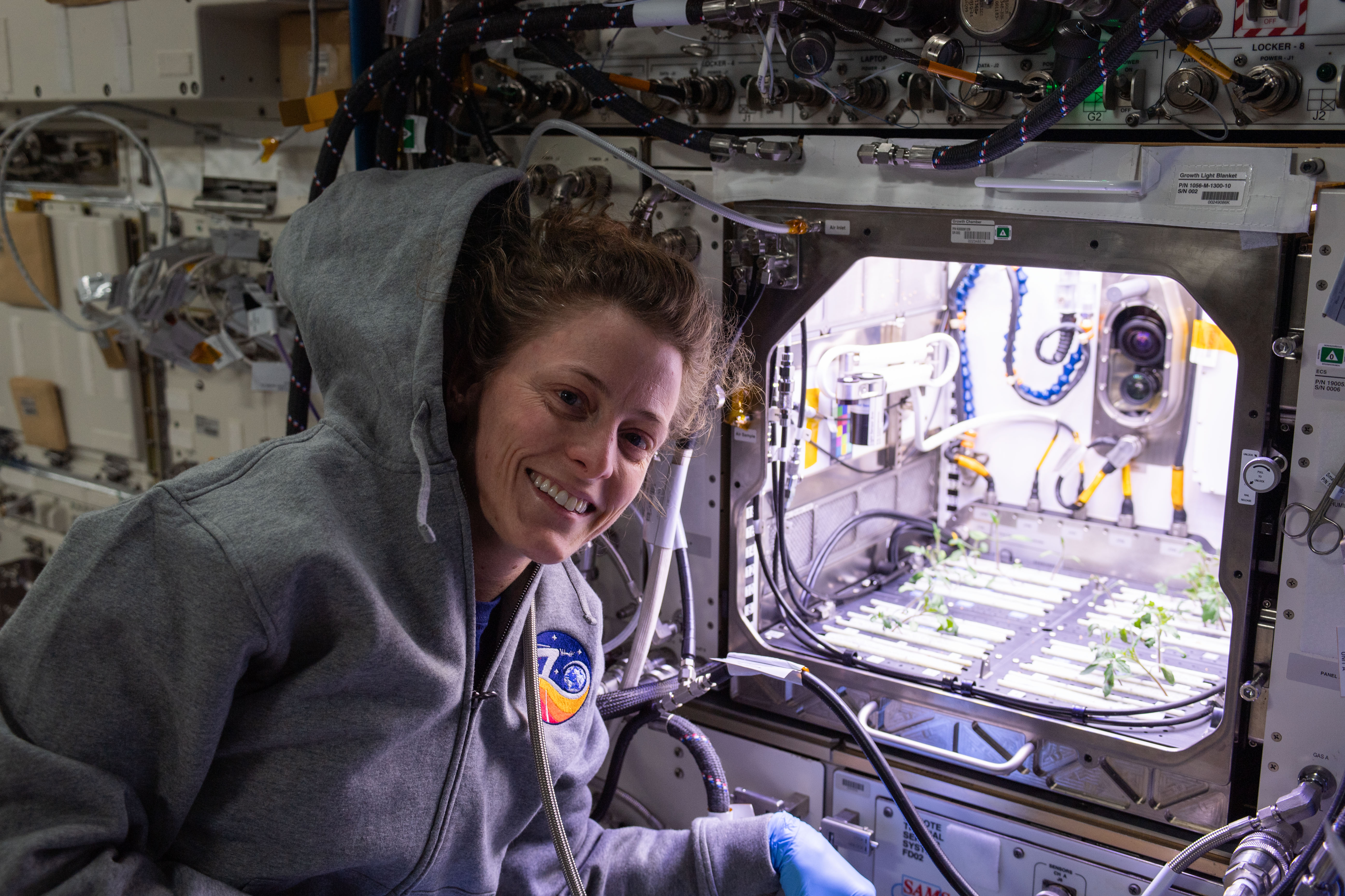 NASA astronaut and Expedition 70 Flight Engineer Loral O'Hara poses in front of the Kibo laboratory module's Advanced Plant Habitat housing tomato plants for an experiment investigating how the plant immune system adapts to spaceflight and how spaceflight affects plant production.
