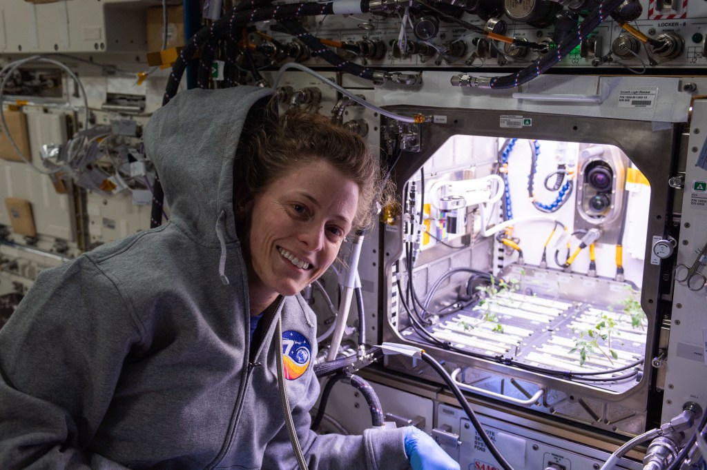 NASA astronaut and Expedition 70 Flight Engineer Loral O'Hara poses in front of the Kibo laboratory module's Advanced Plant Habitat housing tomato plants for an experiment investigating how the plant immune system adapts to spaceflight and how spaceflight affects plant production.