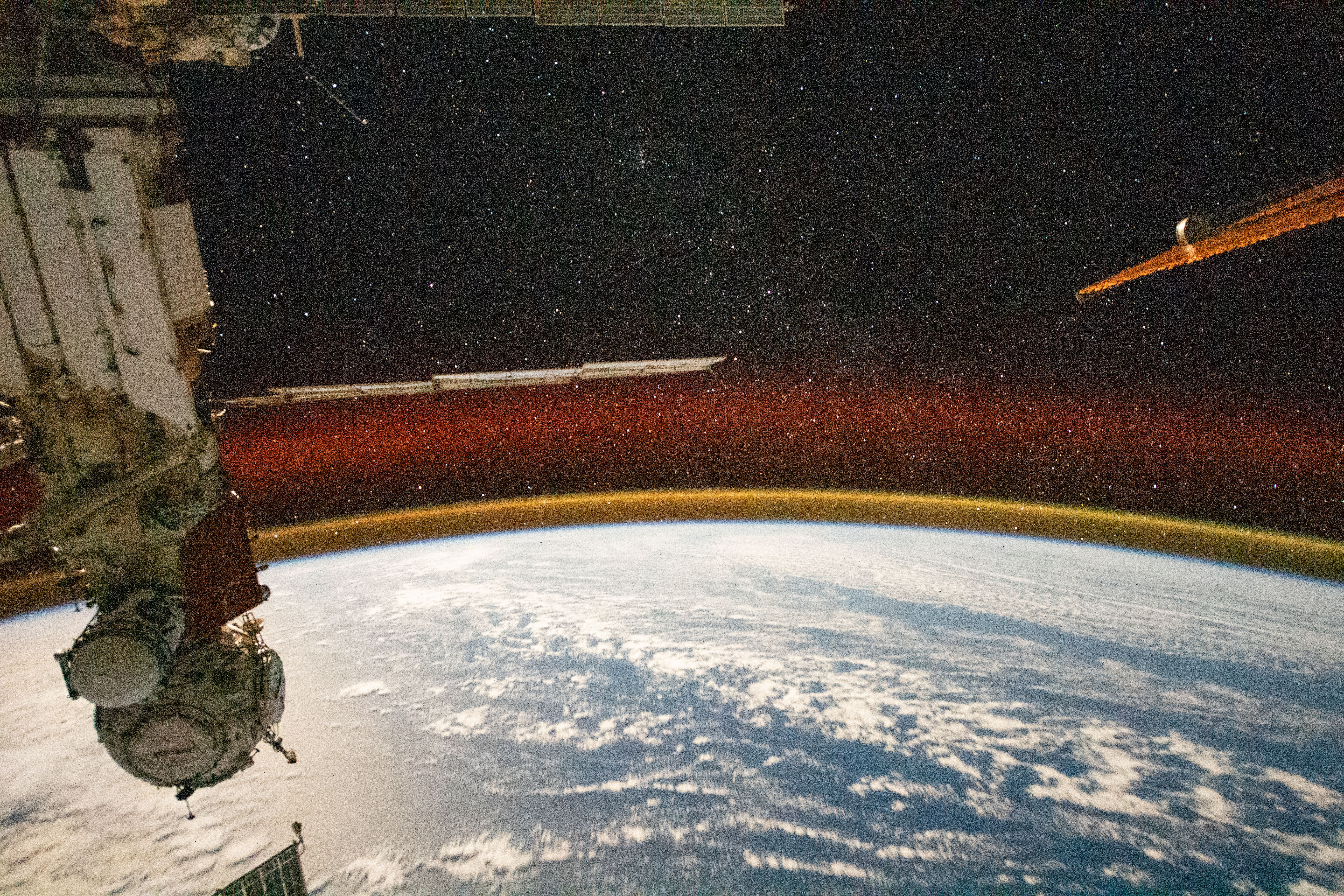 The Pacific Ocean, peppered with bands of white clouds, is seen below a starry sky from the vantage point of the International Space Station. Above the curve of the globe, a well-defined atmospheric glow of yellow-orange is visible, with an additional band of red slightly above. The space station's Nauka science module and Prichal docking module are visible on the left.