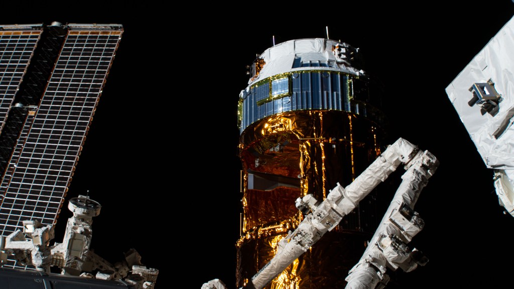 Japan's H-II Transfer Vehicle-9 (HTV-9) is pictured with the elbow of the Canadarm2 robotic arm jutting out in front of it. To the left of the HTV-9 is one of the space station's main solar arrays.