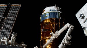 Japan's H-II Transfer Vehicle-9 (HTV-9) is pictured with the elbow of the Canadarm2 robotic arm jutting out in front of it. To the left of the HTV-9 is one of the space station's main solar arrays.