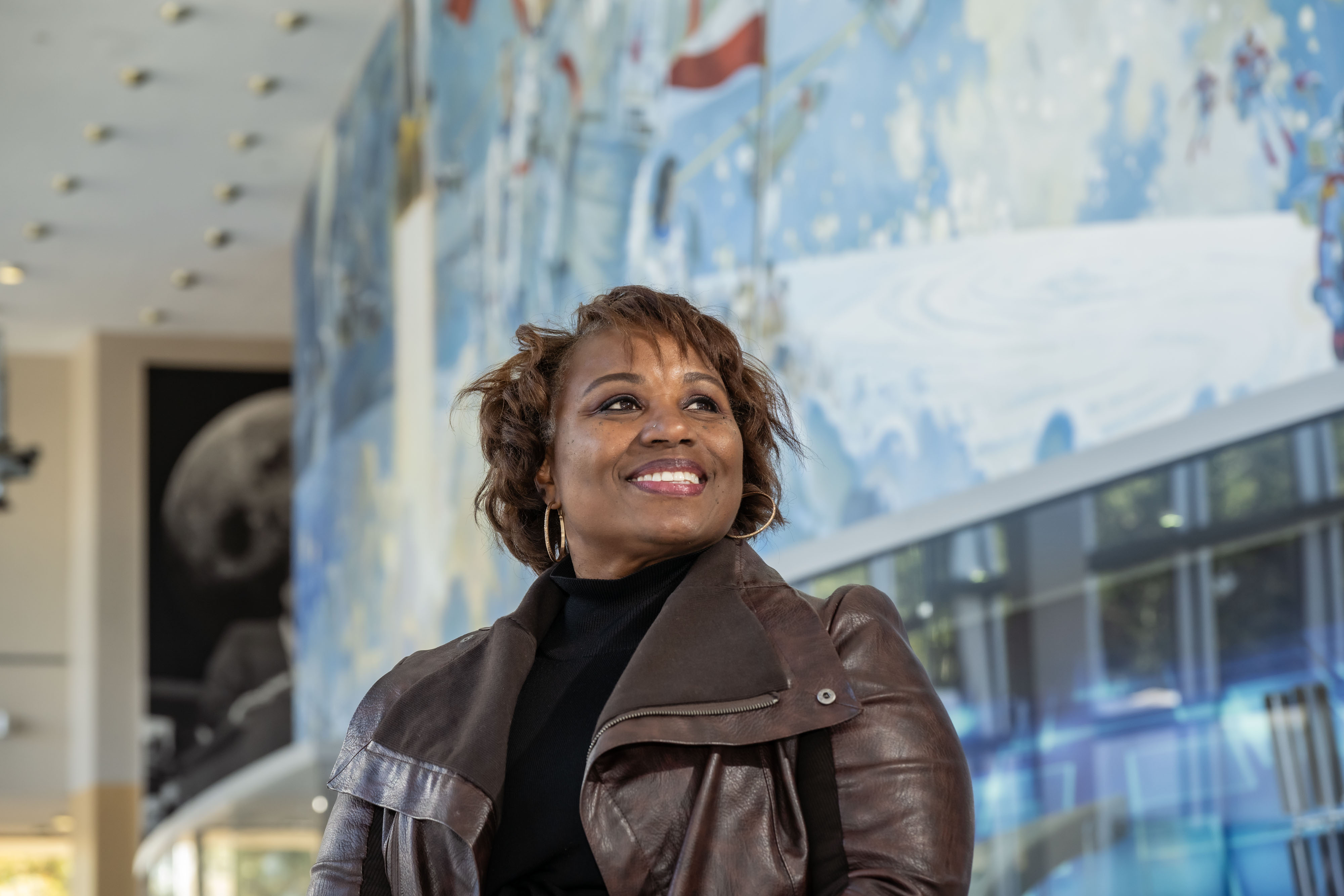 Dr. Camille Alleyne smiles as she looks off in the distance while standing in front of the Robert McCall mural, "Opening the Space Frontier — the Next Giant Step,” in the lobby of Building 2 at NASA's Johnson Space Center.