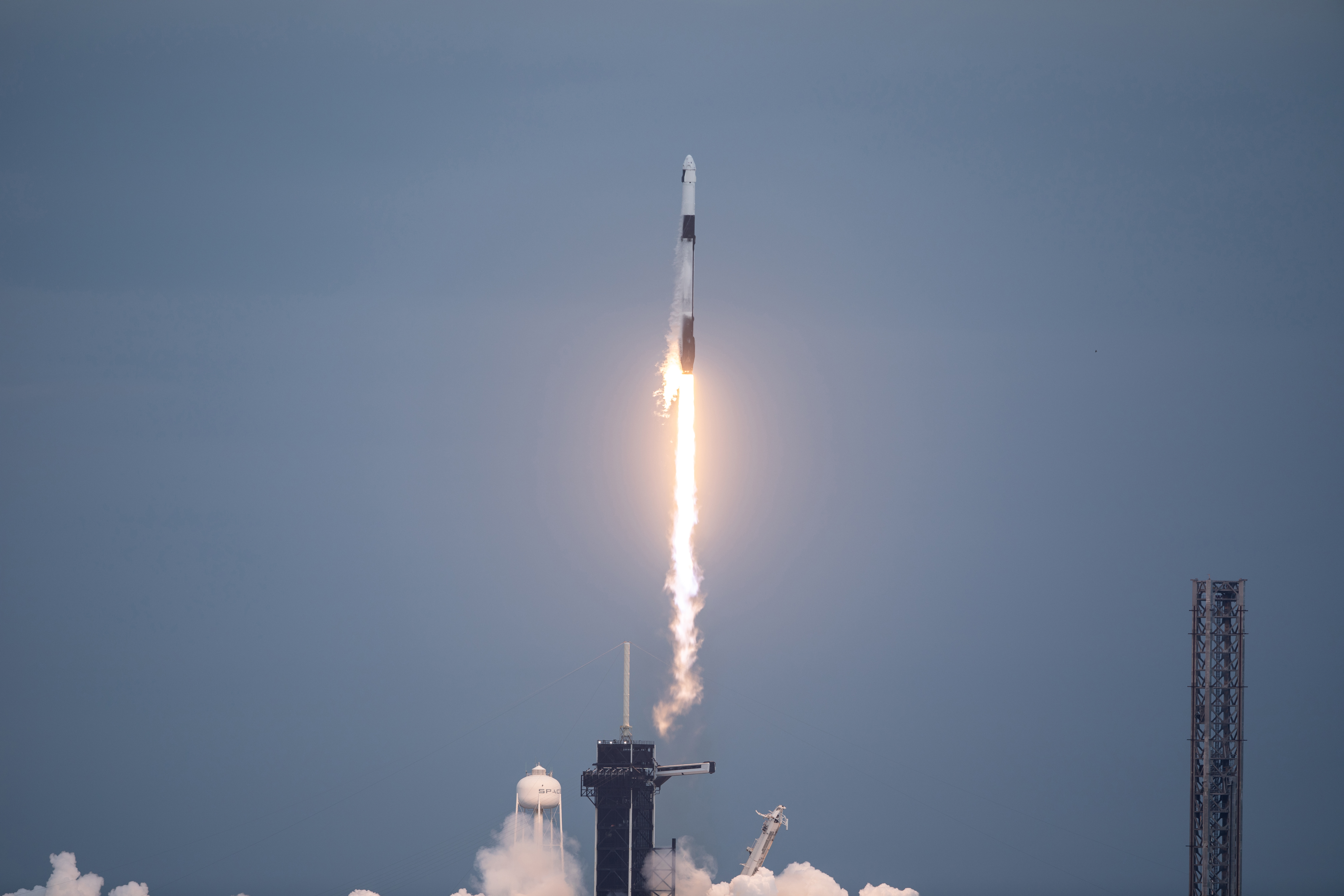 A black and white SpaceX Falcon 9 rocket launches upward through the sky, a bright white-yellow plume trailing behind it.