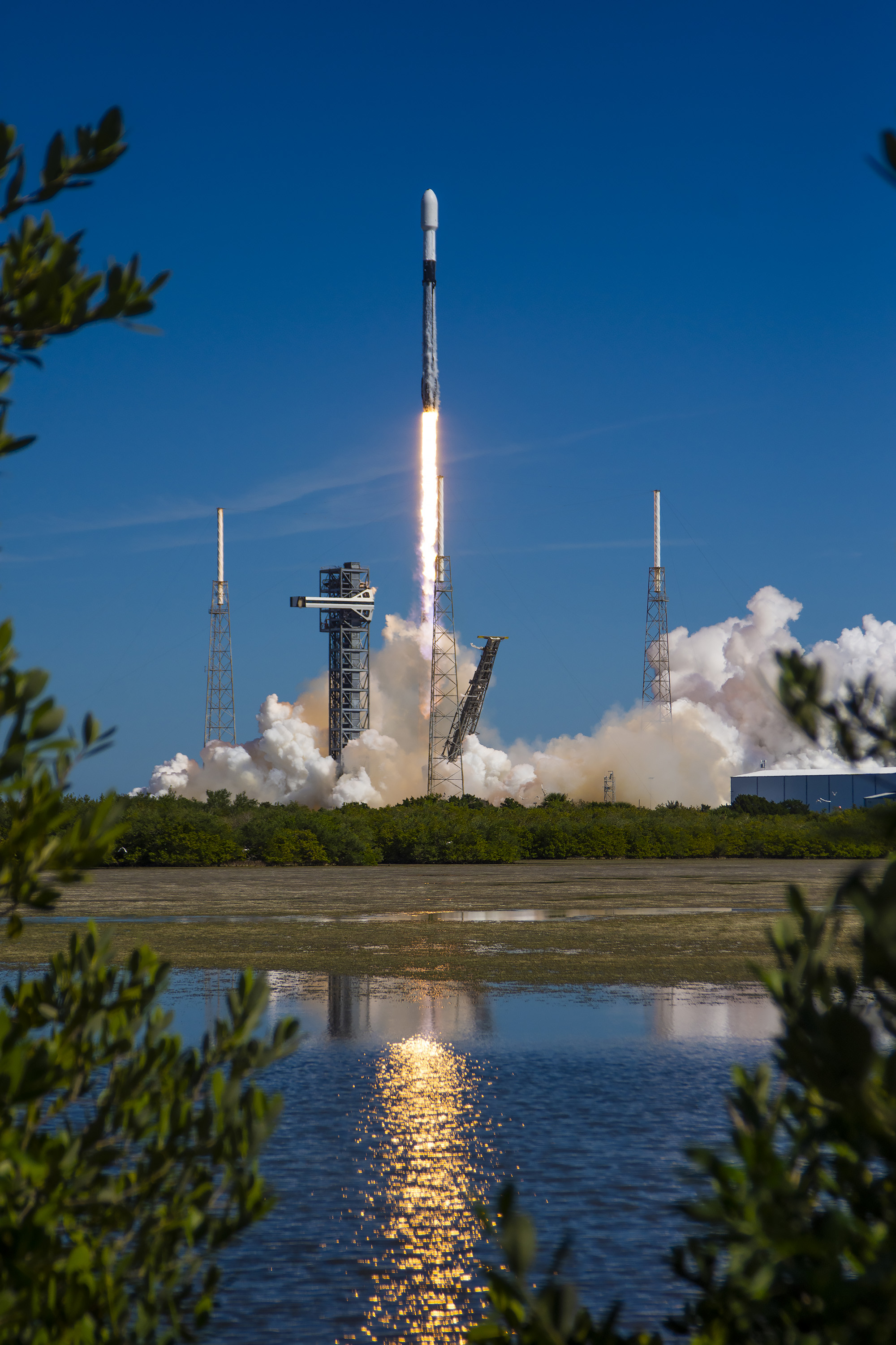 A black and white rocket takes off against the brilliant blue backdrop of the sky. It leaves a trail of fire and billowing clouds of white smoke behind it, which partially obscures some structures around the launchpad. The light reflects on the mirror-like surface of a nearby body of water. The image is delicately framed on the left, right, and bottom by green leaves.