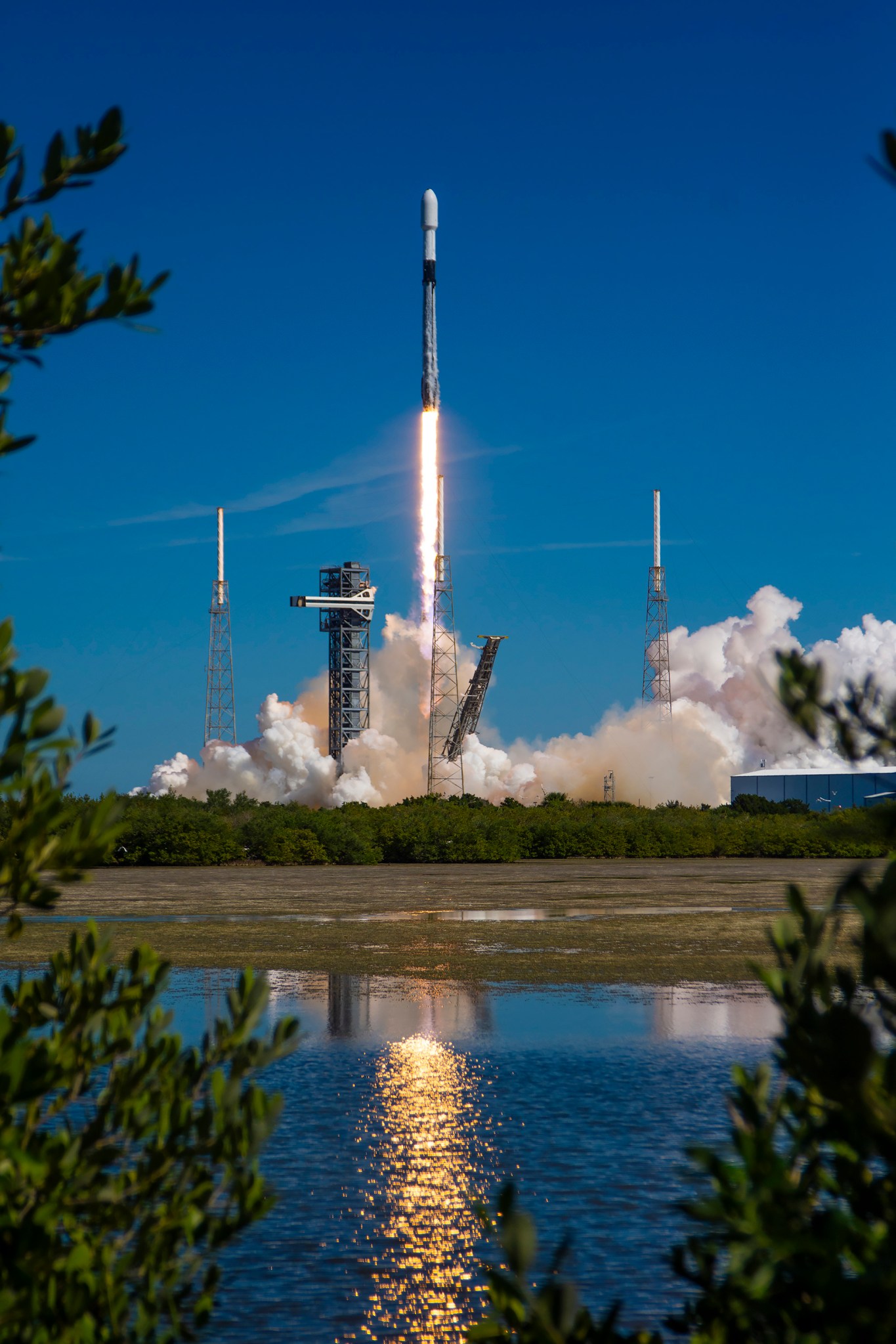 A black and white rocket takes off against the brilliant blue backdrop of the sky. It leaves a trail of fire and billowing clouds of white smoke behind it, which partially obscures some structures around the launchpad. The light reflects on the mirror-like surface of a nearby body of water. The image is delicately framed on the left, right, and bottom by green leaves.