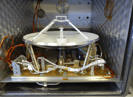 The Polylingual Experimental Terminal is the focus of this photograph. We see a white antenna dish, approximately 0.6-meters in size, facing the ceiling, sitting on a golden platform. Silver wires resembling tinfoil are shown protruding beneath the antenna dish. The terminal sits on top of a grey table inside a white laboratory.  