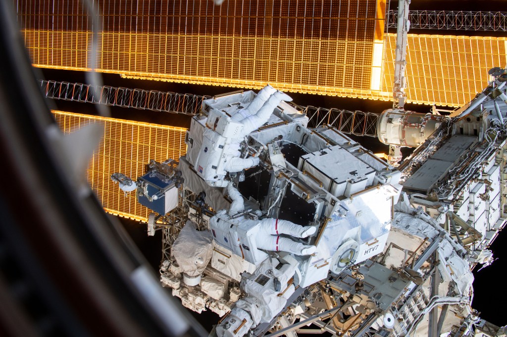 NASA astronauts Nick Hague (suit with no stripes) and Anne McClain (suit with red stripes) work to retrieve batteries and adapter plates from an external pallet during a spacewalk to upgrade the International Space Station's power storage capacity.