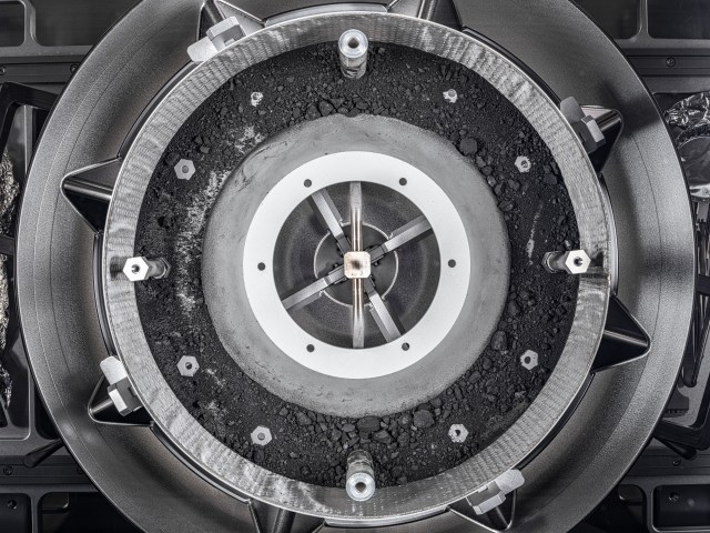 A top-down view of the OSIRIS-REx Touch-and-Go-Sample-Acquisition-Mechanism (TAGSAM) head with the lid removed, revealing the remainder of the asteroid sample inside. The TAGSAM is round, metallic silver, and looks like three concentric circles. The outermost circle has eight triangular metal ridges. The second ring is covered in deep gray, rocky dust particles and small rocks, with silver bolts peeking through the dust. The central circle has three metal bars, crossing in a star pattern.