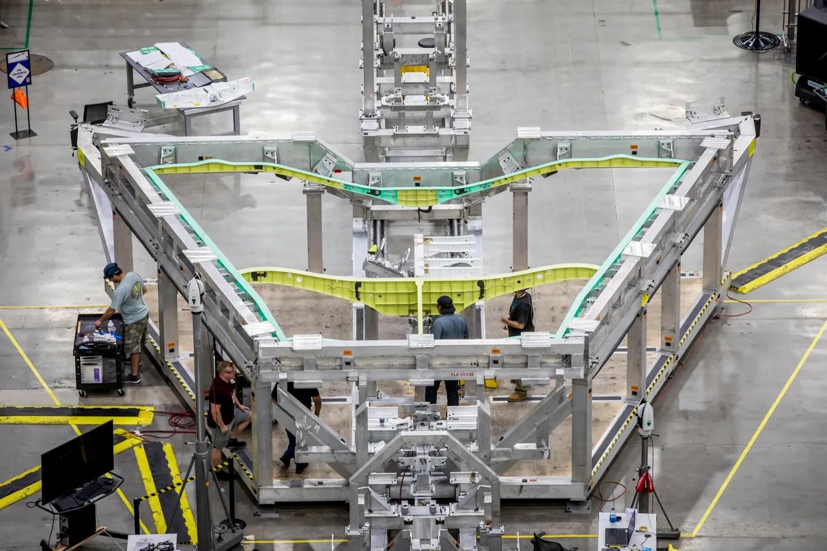 A silver aluminum framework in a roughly triangular shape sits on a clean aircraft factory floor holding green and yellow metal spars that are the ...