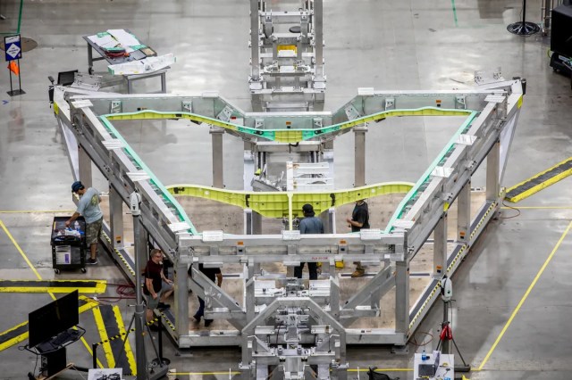 A silver aluminum framework in a roughly triangular shape sits on a clean aircraft factory floor holding green and yellow metal spars that are the first pieces of the wing of NASA's X-59 experimental aircraft.