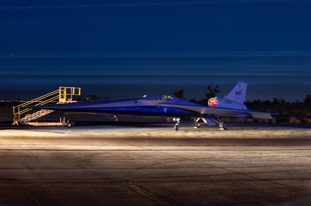 NASA's X-59 airplane is seen parked with the open desert behind it and a row of city lights on part of the horizon.